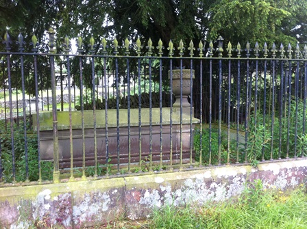 A picture of mid nineteenth century tomb surrounded by an iron fence. In the background is a large tree.