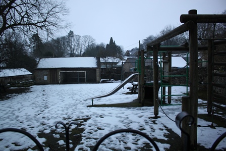 A small children's playground  covered in light snow. There is a climbing frame  and slide to the right and a shed to the left.