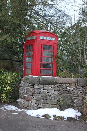 A picture of an red telephone box with a dry stone wall in front of it and trees behind. The sign on the box reads Telephone in capitals. 
