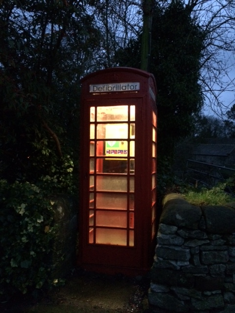 The picture of an old red telephone box at night. The inside of the box is iluminated. The sign on the box reads defibrillator in capitals.