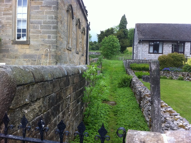 A green path leading about fifty metres to some trees and a tomb. The path runs between an old building to the left and a more modern bungalow to the right.