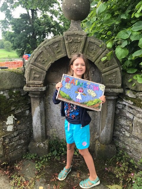A smiling primary school student holding a small well dressing board decorated with petals and featuring the word 'rainbow'.